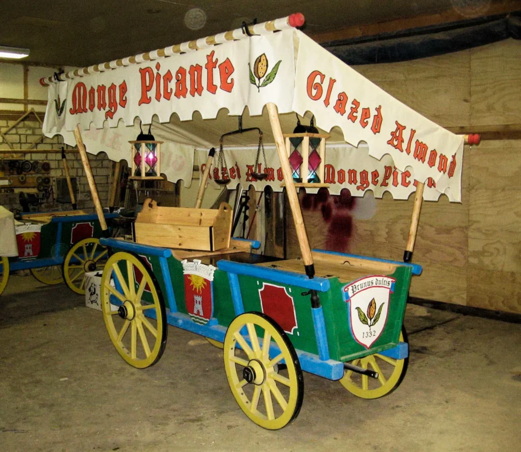 Ein farbenfroher, historischer Marktstand auf Rädern aus Holz mit der Aufschrift „Monge Picante“ und „Glazed Almonds“, ausgestattet mit handgeschmiedeten Lampen und Halterungen in einer Werkstatt.