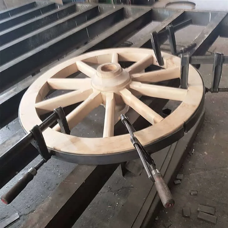 Ein hölzernes Wagenrad liegt auf einem Werkstatttisch aus Metall. Ein flacher Eisenring ist mit mehreren schwarzen Schraubzwingen fest um das Rad gespannt, um die genaue Länge vor dem Schweißen festzulegen.