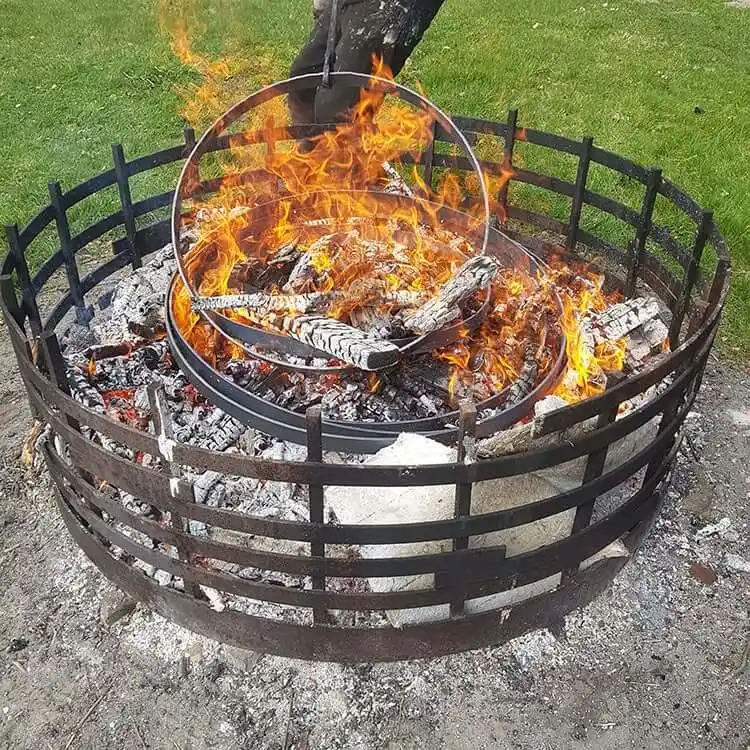 Ein großes, rundes Metallgitter-Lagerfeuer im Freien voller brennendem Holz und glühender Kohlen. Mehrere runde, eiserne Radreifen liegen mitten in den Flammen und der Glut, während einer davon mit einer Schmiedezange bewegt wird.